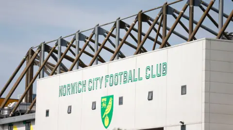 The exterior of a football stadium is shown, with the club's name and crest in large letters on white panels.