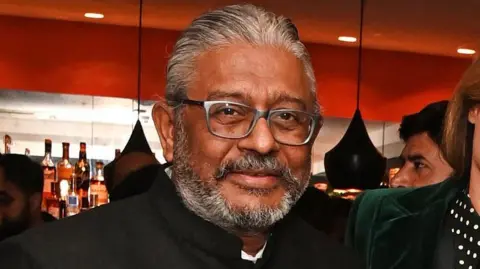 A middle-aged man with a short grey beard, wearing a high-necked suit jacket, looking at the camera.