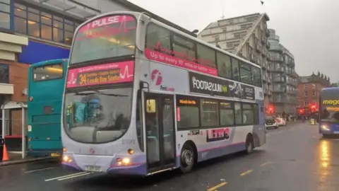 A number 34 bus bound for Leeds Bus Station drives down a wet city centre street.