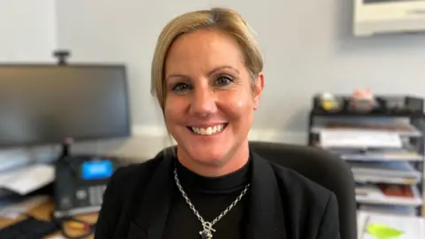 A woman sits in an office at a desk. She has blonde tied back hair and wears a charcoal blazer with a black top underneath and a silver necklace. Behind her is a computer screen and other office equipment. 