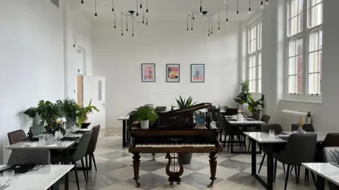 The main dining room of Creeping Thyme which has really tall ceilings with cluster pendant lights with the bulbs on black strings, white walls and wood sash windows. There are marble topped dining tables and a grand piano in the centre of the room with some cocktail artwork on the far wall.