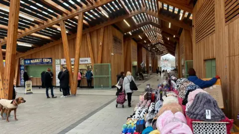 Andrew Turner/BBC Great Yarmouth's market hall, a wooden structure with zinc roof. In the foreground is a market stall selling hats and toys. Beyond that are food stalls where a queue is formed. The picture shows the atrium of the market into the middle distance, with stalls either side, with louvred roof lights throughout.