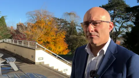 Fred Moroni is bald and is wearing glasses. He is standing outside in front of some stone steps and wooden benches. He is wearing a blue suit jacket and a white shirt with blue stripes