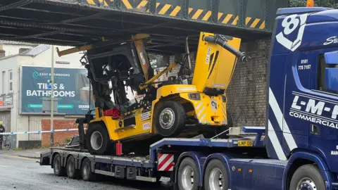 BBC The photo shows a yellow forklift, on the back of a blue lorry trailer, apparently wedged underneath a bridge. Police tape can be seen in the background, as can a billboard on the side of a building.