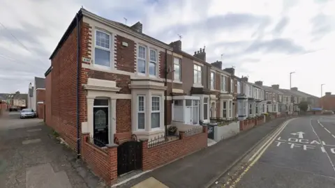 A row of terrace houses on Kent Street in Jarrow. They are made of red brick and have bay windows. A street sign is affixed to the second floor of the first property. 