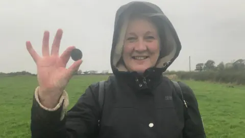 Amanda Parker A woman is smiling while holding what looks like a coin. She is wearing a coat with the hood up and is standing in a field.