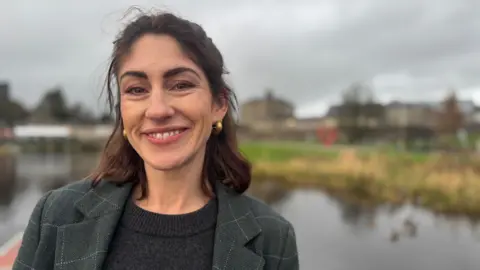 A woman stands by the water. She is wearing a dark green plaid overcoat over a dark woolen jumper. She has shoulder length dark brown hair which is half pinned back. She is smiling and wearing gold earrings. Behind her is a lake and beyond it some land and buildings. 