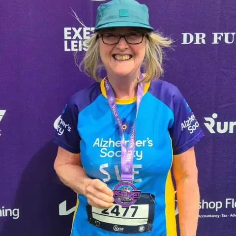 Sue Jackson Sue wearing a cap and a blue and yellow T-shirt. She is standing in front of a sponsor board at the Great West Run. She is wearing dark-rimmed glasses and is smiling broadly at the camera.