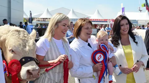 Pacemaker A white cow with a red rosette, Michelle O'Neill holding a rope attached to the cow, a woman holding a baby, and Emma Little-Pengelly.