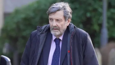 Alan Harris walking. He is wearing a pink top with a blue tie and blazer. He is wearing a black coat. The background is blurred and there is a green bush. 