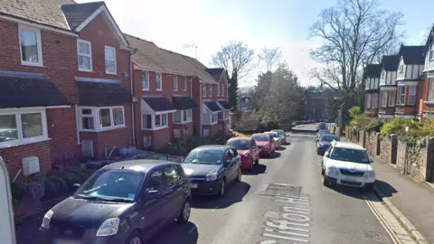 The picture shows a quiet residential street on a sunny day.
On the left side of the scene, there is a line of red‑brick terraced houses with pitched roofs and small front gardens. The houses have white-framed windows and a similar design. Several cars are parked along that side of the road, including a dark grey compact car in the foreground.