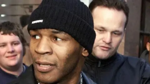 MAN Commercial Protection Mike Tyson in a beanie hat flanked by a fan and a security guard in a black jacket 