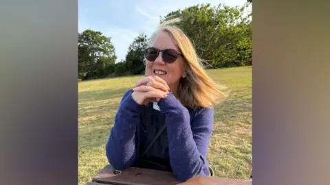 Elizabeth Brown is sitting at a bench and smiling . She is wearing a blue jumper and sunglasses.