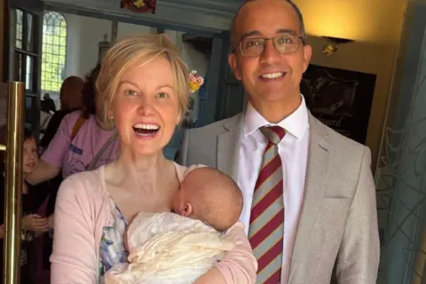 Lucy Turner Lucy has blonde hair which is tied up. She is smiling at the camera. She is wearing a pink blouse. She is holding baby Sandy. Alejandro is smiling at the camera. He has short receding hair and is wearing glasses. He is wearing a suit and stripy tie. You can only see the back of the baby's head. He is wearing a shawl.