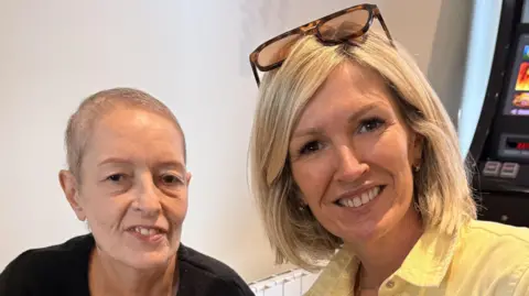 A photo of Rebecca Evans with her sister Jella. Rebecca has blonde hair cut in a bob style and she wears a yellow shirt with sunglasses on her head. Jella next to her wears a black T-shirt. She has a shaved head after cutting her hair prior to cancer treatment. 
