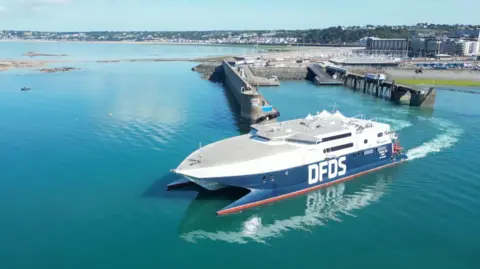 BBC A large blue and white ferry sailing along calm waters. The vessel has a large DFDS logo on its side. The sea water is still and blue. The island is in the background. A pier stretches out into the water.