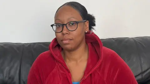 Romesha is wearing a red hooded top and sitting on a black sofa with a painted wall behind her. 