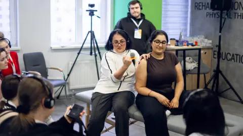 Nataliia Zaslavska Two women are sat on a physiotherapist bench. One is holding a microphone under her arm and has her hand on the shoulder of the other woman as she demonstrates a clinical technique