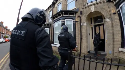 BBC Three police officers, dressed in black uniforms and wearing helmets, approach the front door of a house.
