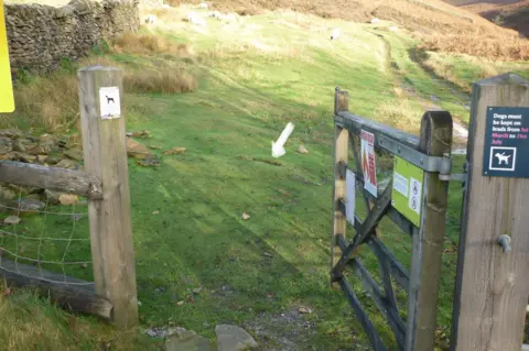 Peak & Northern Footpaths Society An open gate to a field with a white arrow added on the image pointing to a hole in the ground where the sign once stood.