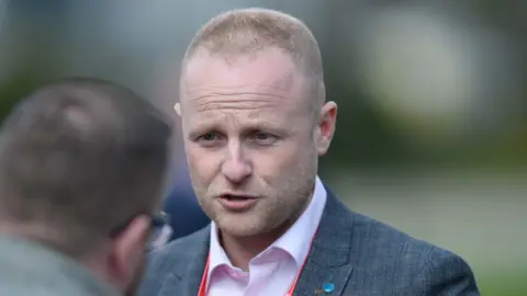 Jamie Bryson has blond short hair, shaved in a number one cut.  He is wearing a grey suit and pink shirt.  He is talking to a man whose head is in shot from behind. 