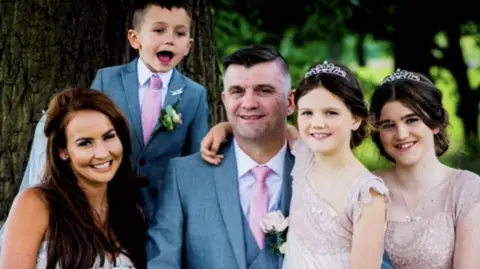 Family photo A wedding photo showing a man and a woman, with their three children, two daughters and a son, smiling next to a tree. 