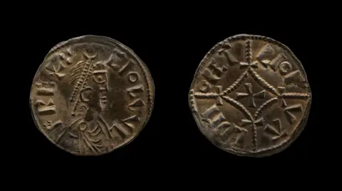 British Museum The photo shows both sides of a small coin, made of silver. It has the profile image of a king on one side and a cross on the other. Both sides include lettering. 