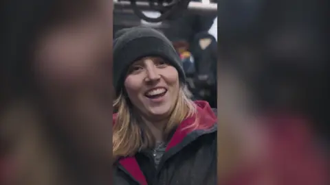 A female farmer in a shed full of cows smiles happily at the camera