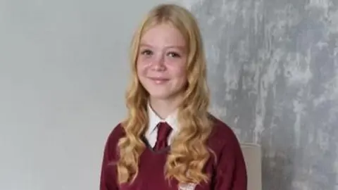 A woman with long blonde hair, wearing a burgundy school uniform is seen smiling at the camera