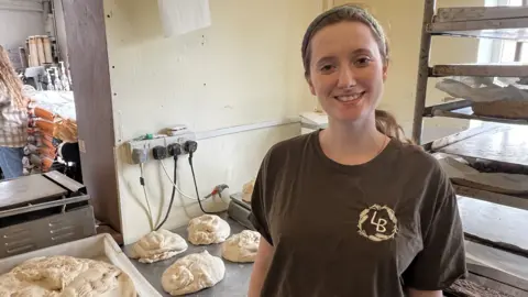 A woman in a bakery with trays of dough.