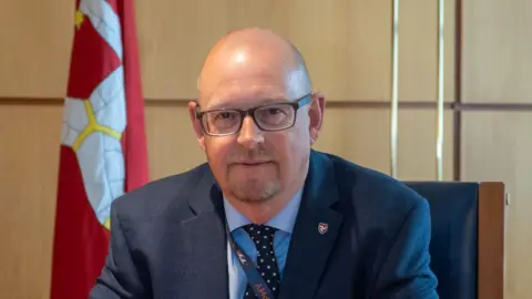 IOM GOV Mark Lewin, a bald man with glasses and grey stubble wears a suit and sits in an office, the Manx flag is in the background.