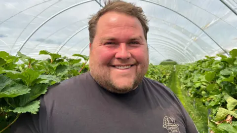 Farmer Joe Hamer smiling inside a polytunnel growing strawberries, wearing a t-shirt branded with his family farm logo