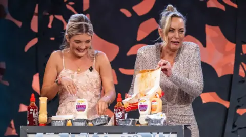 Dominick Walsh Photography Derry Rose Cará Hamilton pictured laughing with host Kathryn Thomas on Monday night's programme. She is wearing a pink dress and Kathryn is wearing a sparkly gown. A number of ingredients are at the table. 