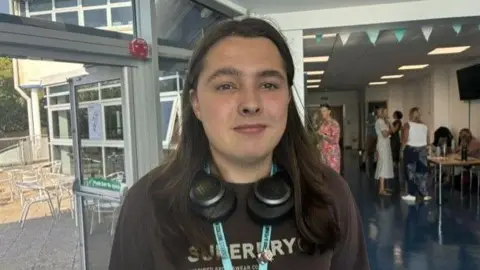 Freddy, a student, has long brown hair and is wearing a brown T-shirt, and some over-ear headphones. He smiles at the camera on results day. 
