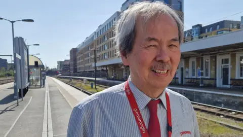 Jeff Tan is standing next to railway tracks on a platform. He has short grey hair and a moustache and is wearing a blue and white striped shirt, red tie and a red lanyard that says "Greater Anglia".