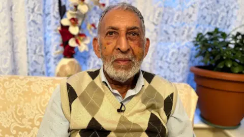 Hukum Dad with grey hair and a beard is wearing a yellow and brown jumper. He is sitting by a window and a plant in a plant pot is on the window sill.