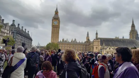 Ollie Conopo/ BBC A group of people gathered in Parliament Square. The Houses of Parliament and Elizabeth Tower are in the background. 