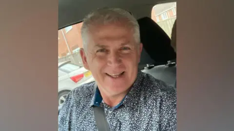 Supplied Steve Howe is smiling in the camera while seating in a car with his seat belt on. He has short white hair and blue eyes. He is wearing a blue shirt with a print of tiny white flowers.