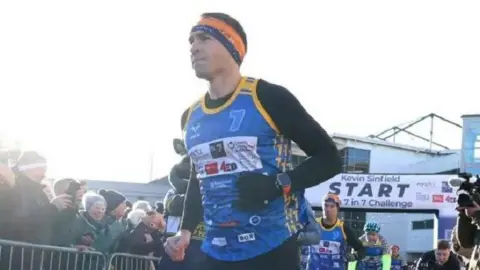 Kevin Sinfield wearing his running gear while completing a previous 7 in 7 challenge. People watch him and other runners behind him.