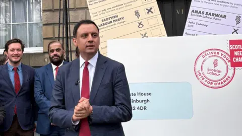 PA Media Anas Sarwar standing in front of a large white envelope and peach and lilac ballot papers outside Bute House in Edinburgh
