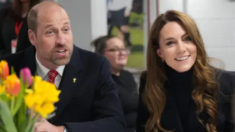 PA Media The Prince and Princess of Wales in the Sir Tasker Watkins Suite in the Principality Stadium. Both aren't looking at the camera. William sits on the left, talking to someone off camera, whilst Kate sits on his right, smiling. In the left corner there are a bouquet of flowers, including daffodils. A woman  wearing glasses can be seen in the background.