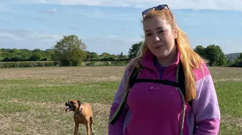 BBC/Tom MacDougall A woman with long ginger hair looks at the camera and smiles. She is wearing a purple jumper and a rucksack. A dog is in the background, standing in a field. It is a sunny day with some cloud in the sky.