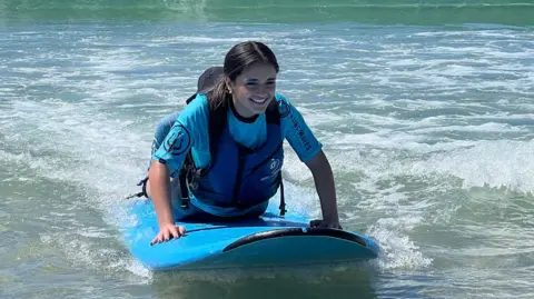 Marah is wearing a blue top and life vest kneels on a blue surfboard in shallow ocean water. Small waves are breaking nearby, and a larger green wave is visible in the background under clear skies.
