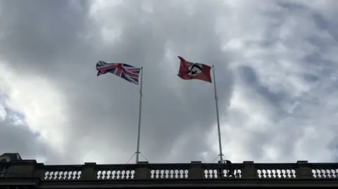 Lara King/BBC Two flags flying in the wind at the top of flag poles on top of a building. One flag is the Union Jack while the other is the flag of Great Grimsby which is red with the town's coat of arms on it.