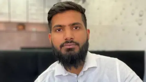 A man wears a white dress shirt and has a microphone attached to his lapel. He has a black beard and dark hair.
