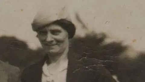 Louisa Dunne in a black and white photograph. She is wearing a hat and a brown jacket with a white shirt. 