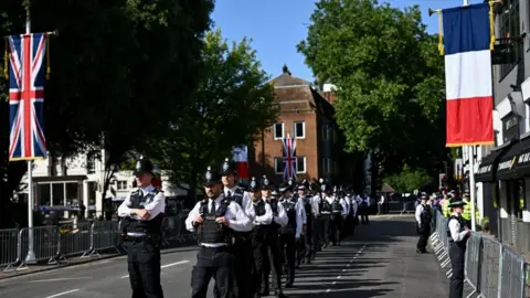 Reuters UK and French flags draped on posts straddle a road which dozens of police officers walk down.