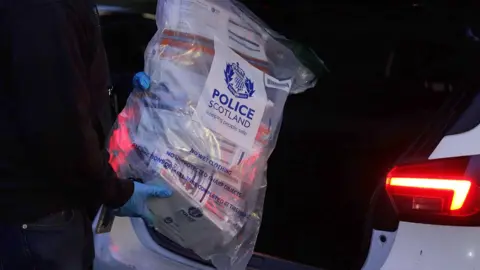 Close up shot of blue latex gloved hands holding a large clear plastic police evidence bag bulging with indistinguishable papers and other contents