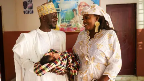 Cem Ozdel / Getty Images A couple - wearing white and gold outfits - gaze at each other white holding a newborn baby in Banjul, The Gambia - 15 February 2025.