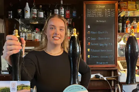 Sophie Landers - a young woman with long blonde hair stands behind a pub bar, smiling, with her hand on a beer pump.